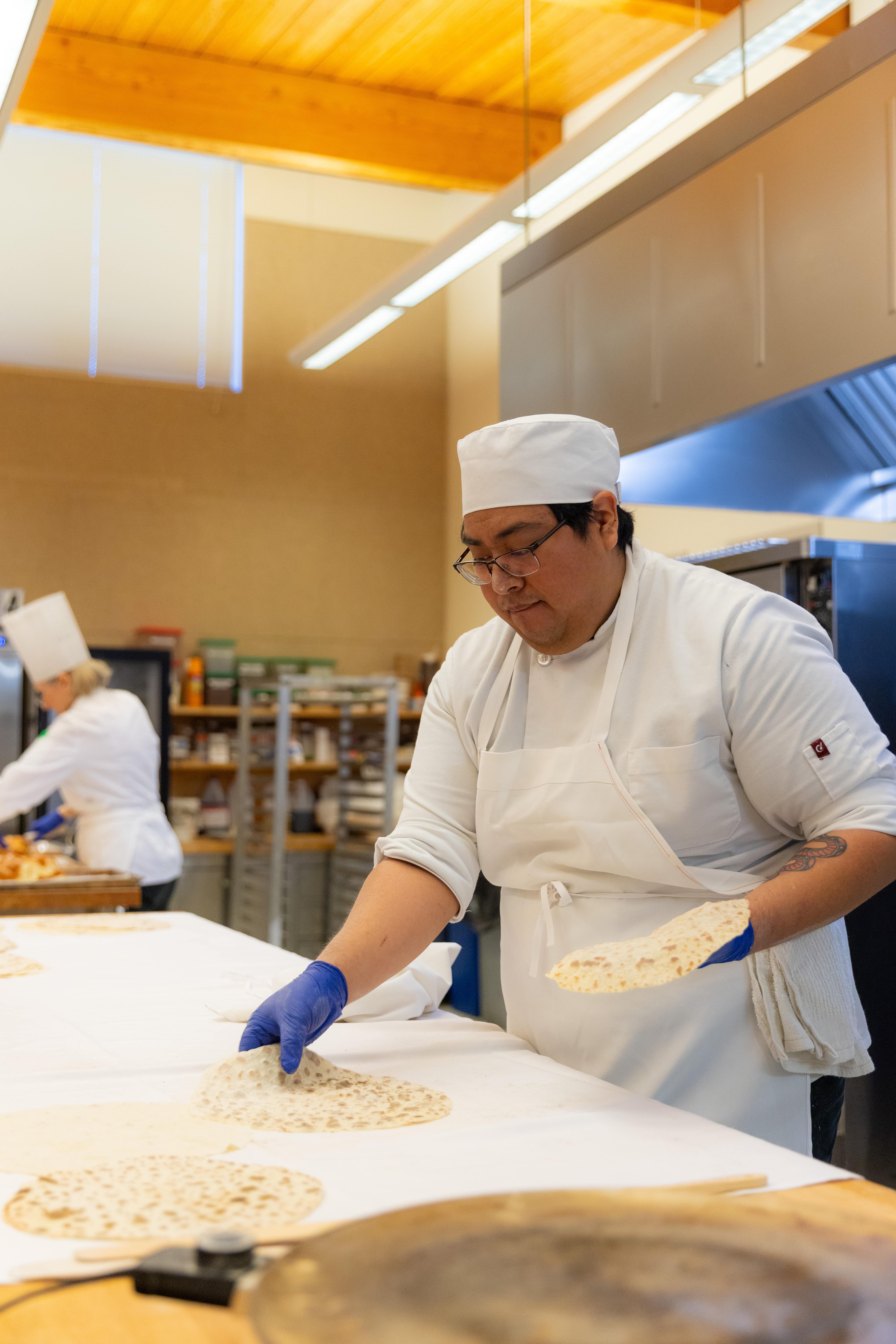 A chef in a white uniform and hat carefully places flatbread on a table in a professional kitchen. Another chef works in the background, creating a busy but focused atmosphere.