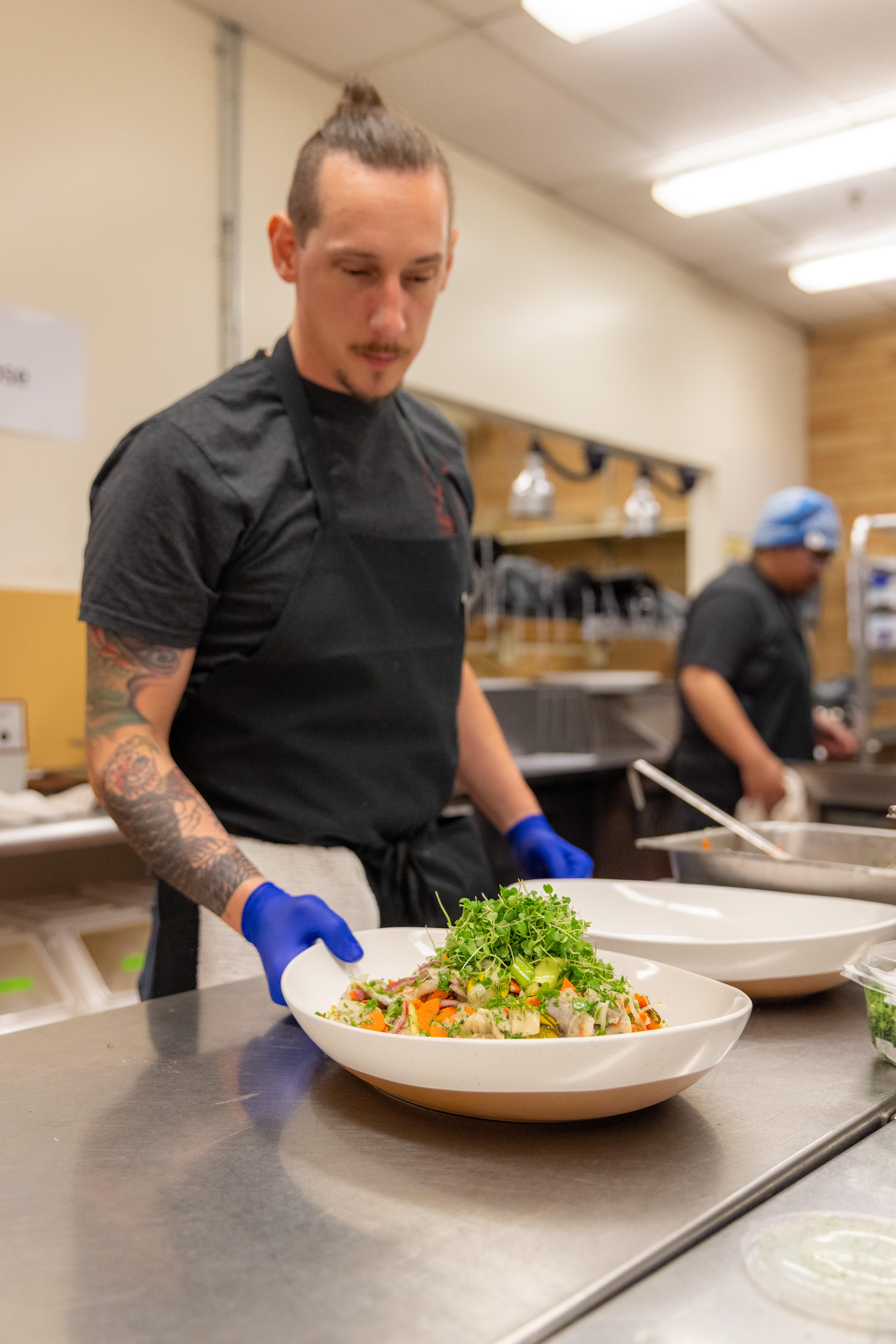 Chef in a commercial kitchen prepares a dish, carefully garnishing it with fresh greens. He wears a black apron and blue gloves, focusing intently.