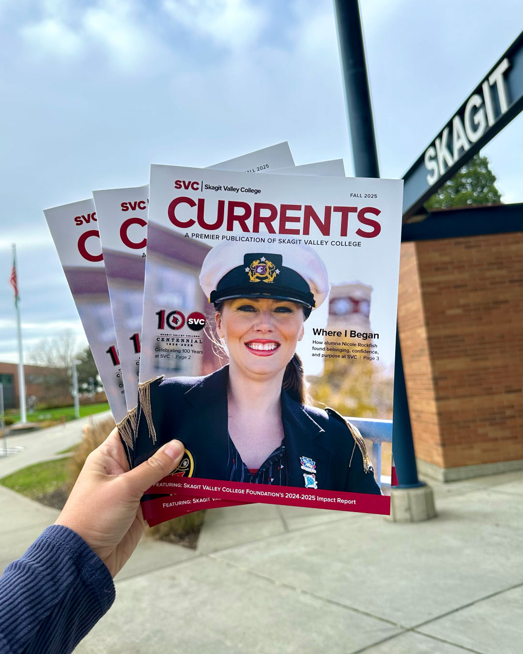A hand holding copies of "Currents," featuring a smiling woman in a naval uniform on the cover. The Skagit Valley College sign is visible in the background.